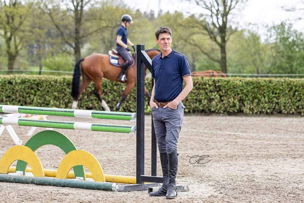 Dieses Bild zeigt einen Mann in Reitkleidung vor einem Springreitschirm, mit einem Pferd im Hintergrund.